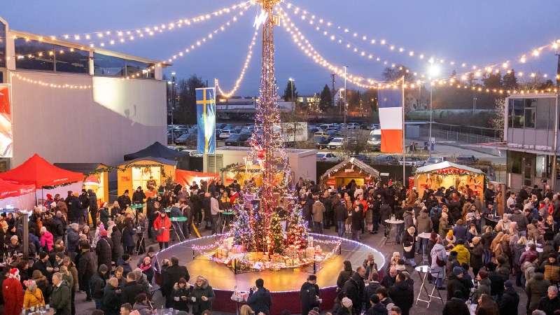 A festive Christmas market scene features a tall, illuminated tree adorned with lights and decorations. Crowds of people in winter attire gather around festive booths, enjoying the lively, cheerful atmosphere. Strings of lights hang overhead, enhancing the holiday mood.