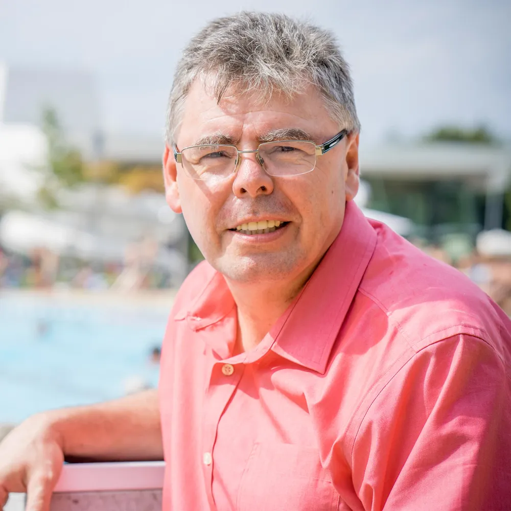 Man in a bright pink shirt smiling outdoors near a swimming pool with blurred people in the background.