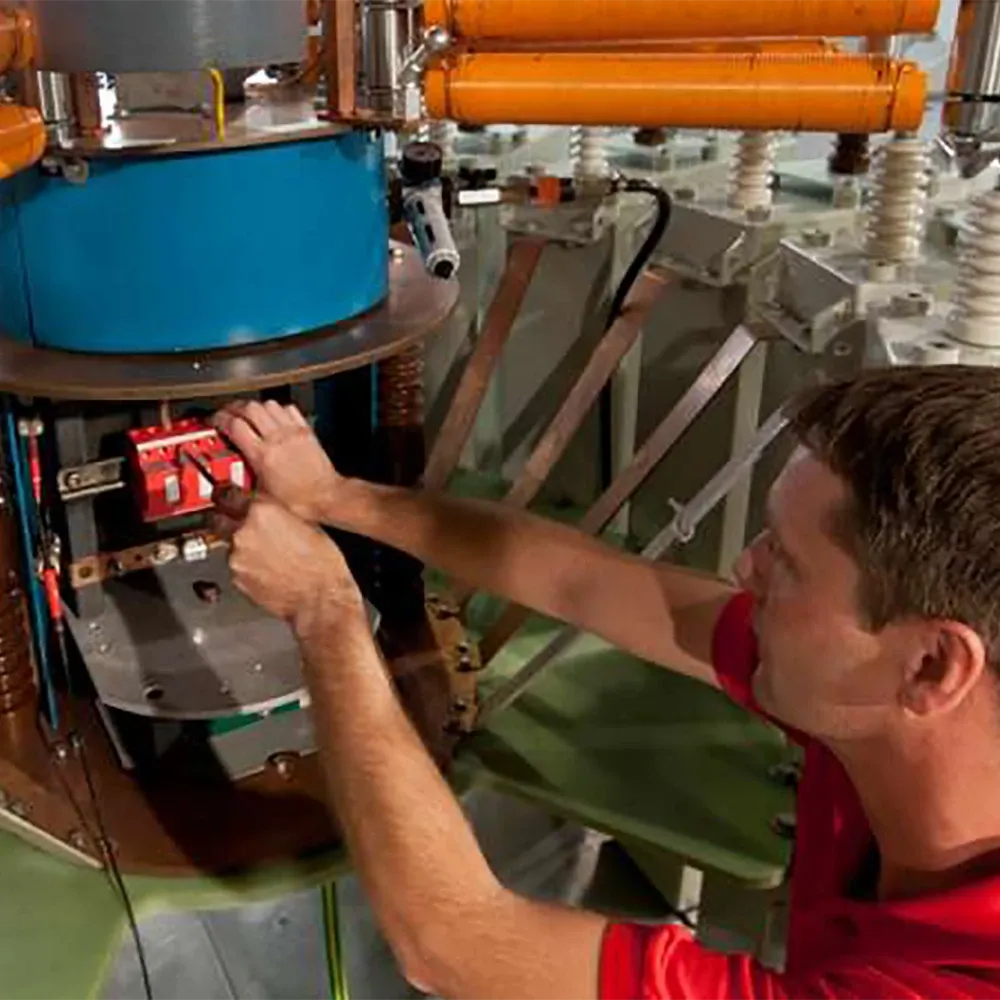 Man adjusting electrical components inside an industrial machine with visible cables, metal parts, and insulated pipes.